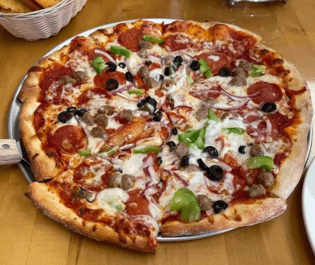 A golden-brown pizza topped with beef, chicken, pepperoni, vegetables, and bubbling cheese fresh out of a wood-fired oven.