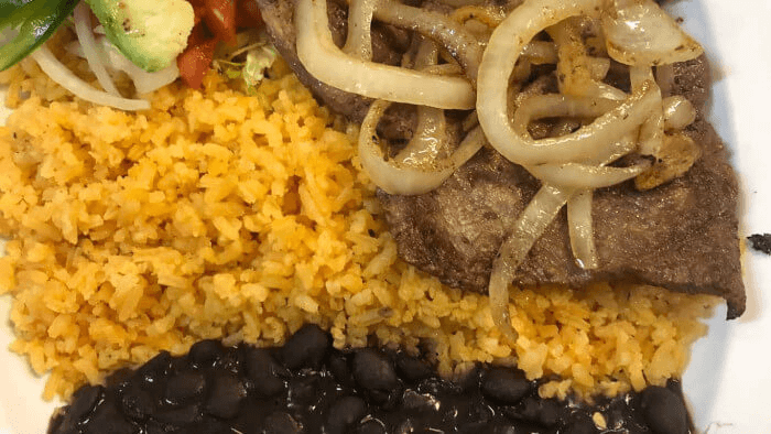 Delicious Tex-Mex plate of sliced steak sautéed with onions, served with Mexican rice, refried beans, and a fresh salad topped with avocado slices.