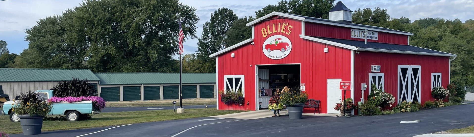 Red barn-style Ollie’s pizza drive-through with white trim, open service window, flowers, and parking lot in a small-town setting.