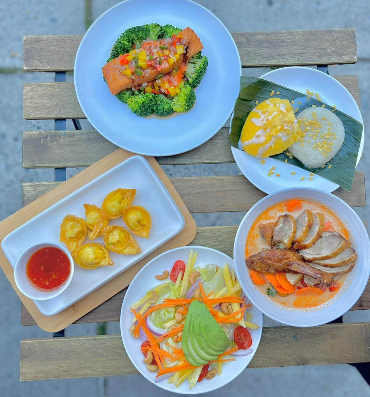 A colorful Thai table spread featuring Shrimp Pad Thai, Papaya Salad, and Curry Puffs with lime wedges, herbs, and chili flakes for garnish.